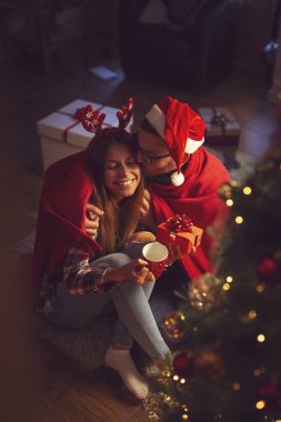 Güzel dekore edilmiş bir Noel ağacının yanında oturan, yumuşak kırmızı battaniyeyle örtülü, sıcak çay içen ve Noel hediyelerini değiş tokuş eden çiftin yüksek açılı görüntüsü.