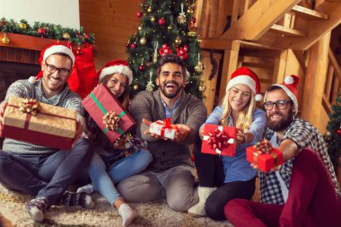 Bir şöminenin yanında oturan bir grup arkadaş ve güzel dekore edilmiş bir Noel ağacı, Noel sabahı eğleniyorlar, Noel hediyelerini değiş tokuş ediyorlar.