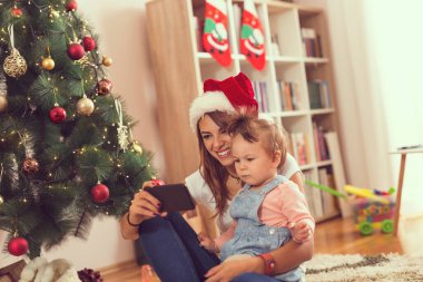 Anne ve kızı bir Noel ağacının yanında yerde oturmuş akıllı bir telefonla çizgi film izliyorlar. Annesine odaklan.