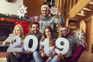 Bir grup genç arkadaş şöminenin yanında oturuyor ve Noel ağacını süslüyor, Noel Baba 'nın şapkalarını takıyor ve ellerinde bir karton kar tanesi ve 2019 numara tutuyor.