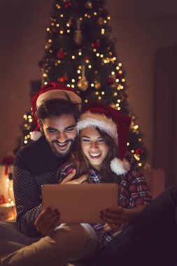 Birbirine aşık bir çift Noel ağacının yanında oturuyor, Noel Baba 'nın şapkalarını takıyor ve tablet bilgisayardaki eski fotoğraflara bakarak eğleniyor. Adama odaklan.