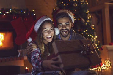 Aşık genç bir çift şöminenin yanında oturuyor ve güzel dekore edilmiş bir Noel ağacı, hediyeleri açıyor ve Noel arifesinde eğleniyorlar. Kıza odaklan.