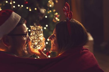 Güzel dekore edilmiş bir Noel ağacının yanında oturan aşık çift, yumuşak kırmızı battaniyeyle kaplanmış, sarılıyor ve Noel arifesi büyüsünün tadını çıkarıyor. 