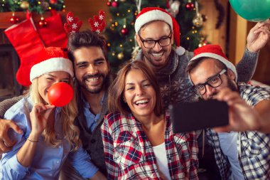 Bir grup arkadaş şöminenin ve Noel ağacının yanında oturmuş Noel sabahı eğleniyor, renkli balonlar üflüyor ve selfie çekiyorlar.