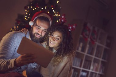 Birbirine aşık bir çift Noel ağacının yanında oturuyor, Noel Baba şapkası takıyor ve tablet bilgisayardaki eski fotoğraflara bakarak eğleniyor..