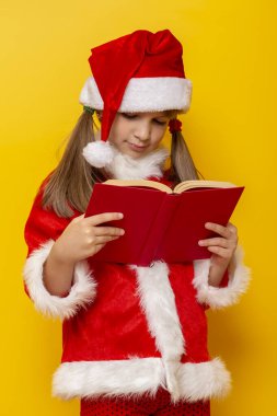 Noel Baba kostümü giymiş güzel bir kız kitap okuyor, sarı renkli arka planda izole edilmiş.