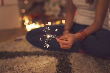 Kadın elinin ön planda Noel ışıkları ve sırtında Noel ağacı olan bir maytap tutmasının ayrıntıları.