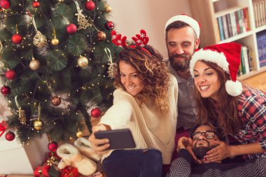 Bir grup genç arkadaş, Noel Baba şapkası takıp selfie çekerek güzel dekore edilmiş bir Noel ağacının yanında yerde oturuyorlar. Telefonlu kıza odaklan.