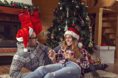 Birbirine aşık güzel bir çift, Noel Baba şapkası takıyor, güzel dekore edilmiş bir Noel ağacının yanında uzanıyor ve şampanya kadehleriyle kadeh kaldırıyor.