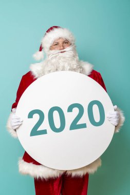 Mutlu bir Noel Baba 'nın portresi, üzerinde 2020 rakamları bulunan bir karton daire tutuyor. Kış satış sezonunu ya da yeni yıl partisi davetiyesini temsil ediyor. Renkli arka planda izole edilmiş.