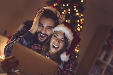 Birbirine aşık bir çift Noel ağacının yanında oturuyor, Noel Baba 'nın şapkalarını takıyor ve tablet bilgisayardaki eski fotoğraflara bakarak eğleniyor. Kıza odaklan.