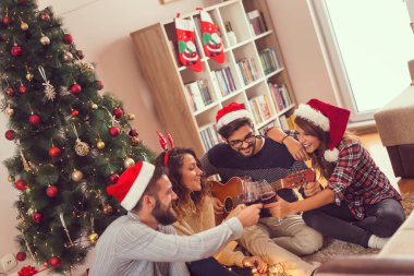 Bir grup arkadaş güzel dekore edilmiş Noel ağacının yanında oturmuş gitar çalıyor, Noel şarkıları söylüyor ve şarap kadehleriyle kadeh kaldırıyorlar.