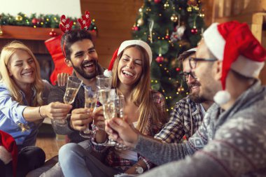 Bir grup arkadaş yerde güzel dekore edilmiş bir Noel ağacının yanında oturmuş şampanya kadehleriyle kadeh kaldırıyorlar. Ortadaki kıza odaklan.