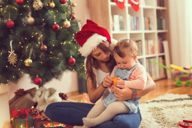 Genç bir anne bebeğini kucağında tutuyor, güzel dekore edilmiş bir Noel ağacının yanında yerde oturuyor ve mandalina soyuyor.    