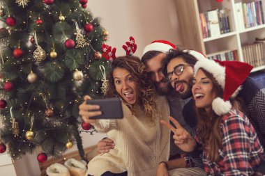 Güzel dekore edilmiş bir Noel ağacının yanında Noel Baba şapkası takıp selfie çeken bir grup genç arkadaş. Soldaki çifte odaklan.