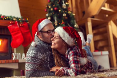 Güzel, birbirine aşık genç bir çift Noel Baba şapkaları takıyor, şöminenin yanında yerde yatıyor ve güzel dekore edilmiş bir Noel ağacı ve bir Noel sabahı öpüşüyor.