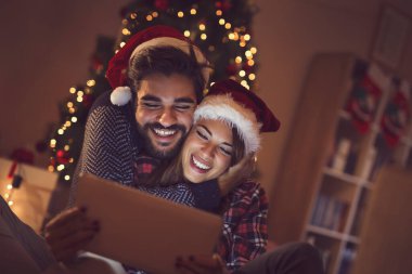 Birbirine aşık bir çift Noel ağacının yanında oturuyor, Noel Baba şapkası takıyor ve tablet bilgisayardaki eski fotoğraflara bakarak eğleniyor.
