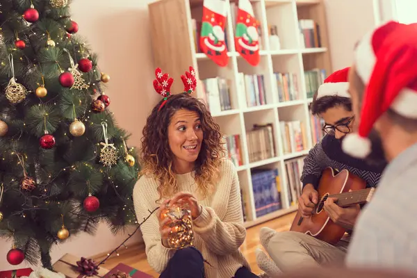 Bir grup arkadaş güzel dekore edilmiş bir Noel ağacının yanında oturmuş gitar çalıyor, Noel şarkıları söylüyor ve Noel günü eğleniyorlar.