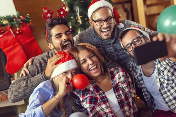 Bir grup arkadaş şöminenin ve Noel ağacının yanında oturup Noel sabahı eğleniyorlar renkli balonlar şişirip evi dekore ederken selfie çekiyorlar.