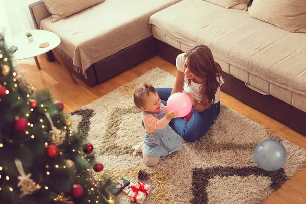 Güzel bir anne ve kızın Noel ağacının yanında oturmuş, Noel sabahı balon şişirirken yüksek açılı görüntüsü.