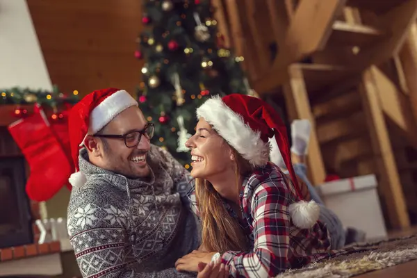 Güzel, birbirine aşık genç bir çift Noel Baba şapkaları takıyor, şöminenin yanında yere uzanıyor ve güzel dekore edilmiş bir Noel ağacı Noel sabahı sarılıyorlar.