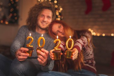 Birbirine aşık bir çift Noel ağacının yanında oturuyor, yeni yıl arifesinde evde eğleniyor, ellerinde 2020 numaralı mumlar şeklinde yanan çikolatalı kekler tutuyorlar.