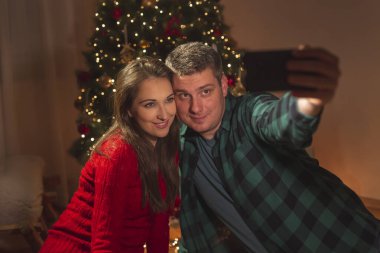 Güzel, genç ve aşık bir çift güzel dekore edilmiş Noel ağacıyla selfie çekiyor, evde Noel 'i kutlarken eğleniyorlar.