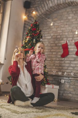 Mutlu anne ve kız Noel ağacının yanında oturuyor, eğleniyor, eğleniyor ve eğleniyorlar.
