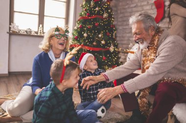 Mutlu bir aile Noel 'i evde kutluyor, çocuklar dedeleriyle güzelce süslenmiş Noel ağacıyla oynuyorlar.