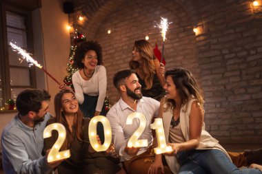 Yeni yıl partisinde eğlenen bir grup genç yeni yılı temsil eden 2021 numaralı aydınlatma numaralarını tutuyorlar ve gece yarısı havai fişeklerle el sallıyorlar.