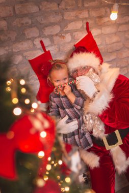 Noel Arifesi 'nin tadını çıkaran küçük güzel bir kız Noel Baba kılığında babasıyla oynarken elinde şeker kamışı bıyığıyla güzelce süslenmiş Noel ağacının yanında duruyor.