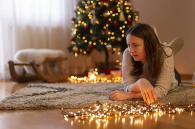 Genç bir kadın kış tatili sezonunda evde vakit geçirir, oturma odasında yere uzanır, güzel dekore edilmiş bir Noel ağacının yanında Noel ışıklarını düzenler.