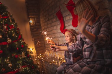 Güzel anne ve kız güzel dekore edilmiş bir Noel ağacının yanında oturmuş, elinde maytap tutarak Noel arifesinde evde eğleniyorlar.
