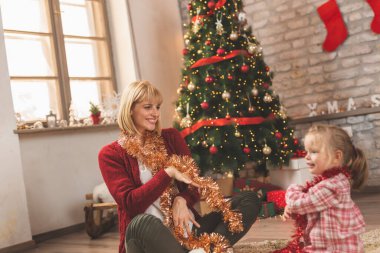 Neşeli anne ve kız Noel ağacının yanında oturmuş, Noel günü evde oynarken eğleniyorlar.