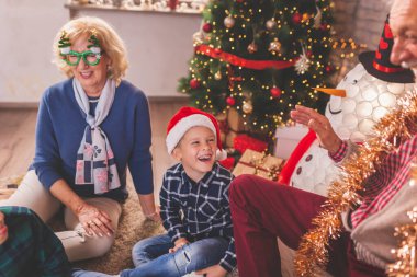 Mutlu bir aile Noel 'i evde kutluyor, çocuklar dedeleriyle güzelce süslenmiş Noel ağacıyla oynuyorlar.