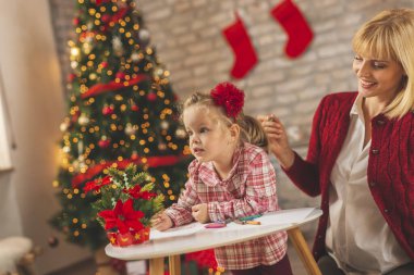 Anne ve kızı Noel günü evde eğleniyor, güzelce süslenmiş Noel ağacının yanında oturuyor ve Noel Baba 'ya mektup yazıyor ve resim çiziyor.