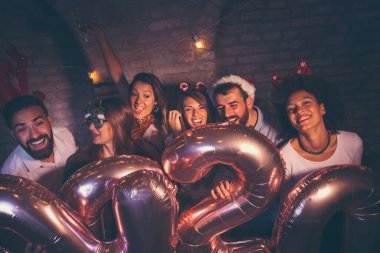 Bir grup genç arkadaş yeni yıl partisinde eğleniyor, ellerinde 2020 numara şeklinde balonlar var ve gece yarısı geri sayımında şampanya içiyorlar.