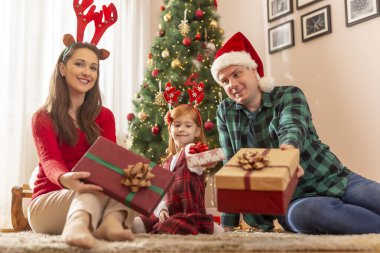 Anne, baba ve kız güzelce süslenmiş Noel ağacının yanında oturmuş, evde Noel hediyelerini verirken eğleniyorlar.