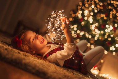 Güzel küçük kız yere uzanmış güzel süslenmiş Noel ağacının yanında, elinde bir demet Noel ışığı ve gülümsüyor.