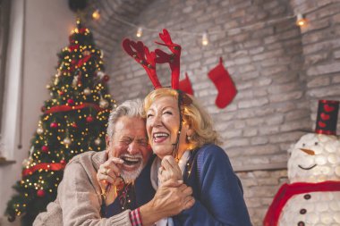 Güzel dekore edilmiş bir Noel ağacının yanında oturan mutlu yaşlı çift Noel ışıklarını tutuyor ve Noel günü evi dekore ederken eğleniyorlar.