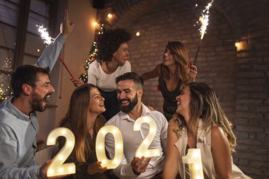 Yeni yıl partisinde eğlenen bir grup genç yeni yılı temsil eden 2021 numaralı aydınlatma numaralarını tutuyorlar ve gece yarısı havai fişeklerle el sallıyorlar.