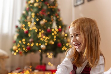 Güzel küçük kız Noel günü evde oynarken eğleniyor, güzelce süslenmiş Noel ağacının yanında oturuyor ve gülümsüyor.