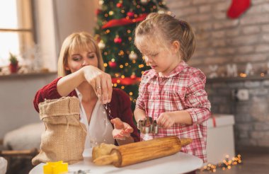 Anne ve kız Noel ağacının yanında oturmuş zencefilli kurabiye yapıyor ve eğleniyorlar.