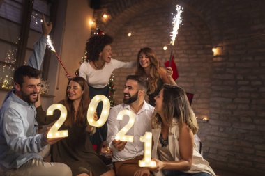Yeni yıl partisinde eğlenen bir grup genç yeni yılı temsil eden 2021 numaralı aydınlatma numaralarını tutuyorlar ve gece yarısı havai fişeklerle el sallıyorlar.