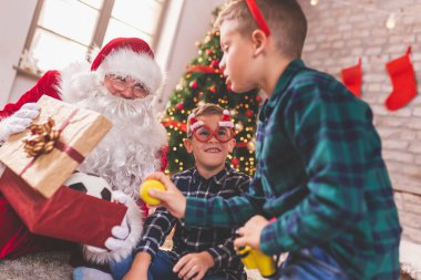 Noel Baba 'yla Noel ağacının yanında oturan iki küçük çocuk Noel hediyelerini açıyor ve evde yeni oyuncaklarıyla eğleniyorlar.