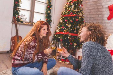 Genç ve güzel bir çift evde Noel 'i kutlarken kadeh kaldırıyor ve şarap içiyor, Noel ağacının yanında oturuyor ve kış tatilinin tadını çıkarıyor.