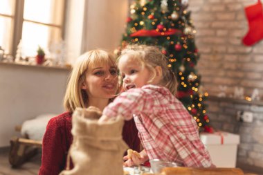 Anne ve kız Noel ağacının yanında oturmuş zencefilli kurabiye yapıyor ve eğleniyorlar.