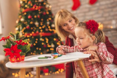 Anne ve kızı Noel günü evde eğleniyor, güzelce süslenmiş Noel ağacının yanında oturuyor ve pastel boyalarla çiziyor, Noel Baba için bir mektup süslüyor.