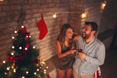 Güzel dekore edilmiş Noel ağacının yanında duran, şarap içen ve Noel 'i evde kutlarken eğlenen güzel genç çiftin yüksek açılı görüntüsü.