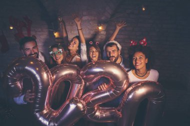 Bir grup genç arkadaş yeni yıl partisinde eğleniyor, ellerinde 2020 numara şeklinde balonlar var ve gece yarısı geri sayımında şampanya içiyorlar.